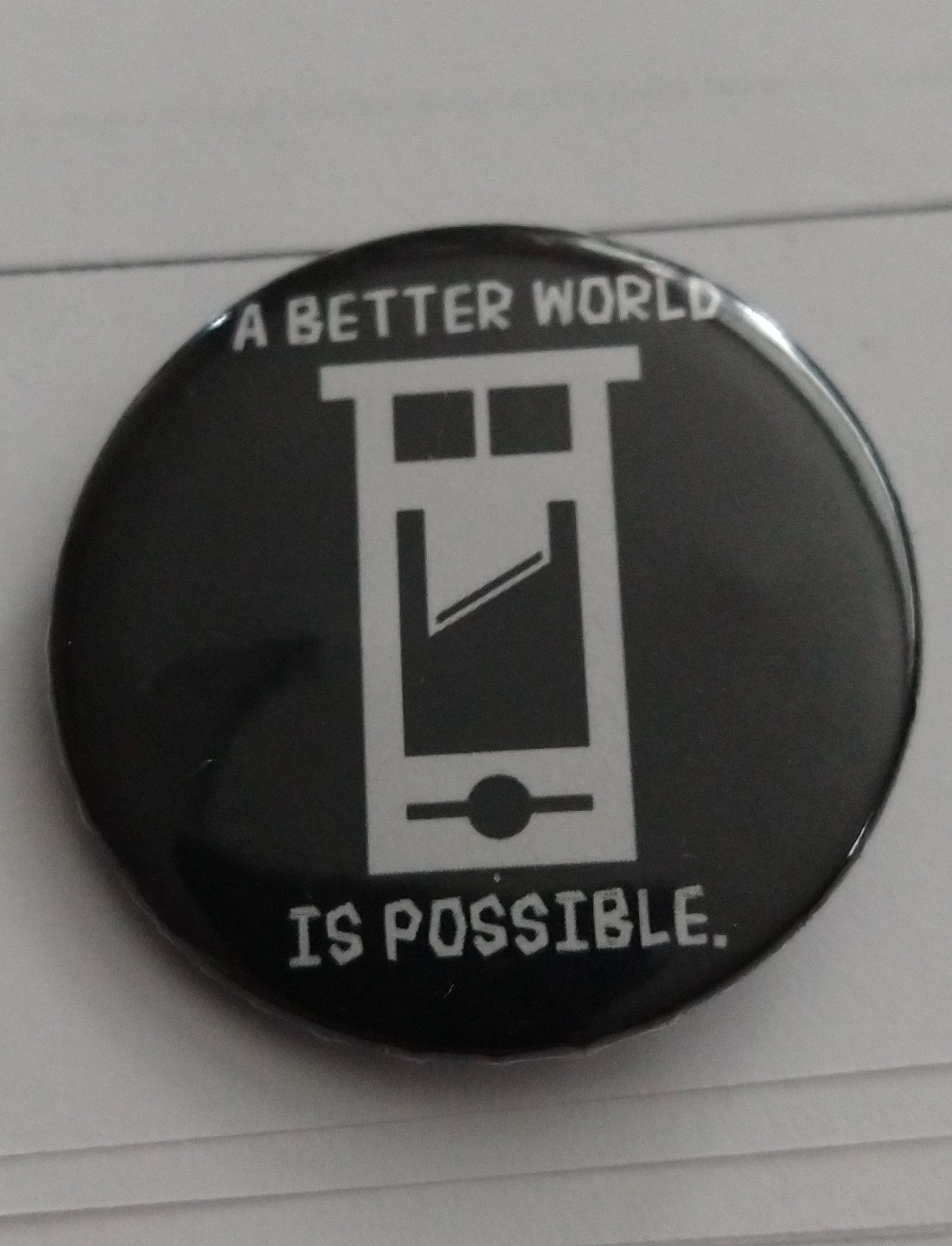 A picture of a black circular pin with a white guillotine in the center of the pin. The inscription above and below the guillotine reads "A Better World Is Possible"