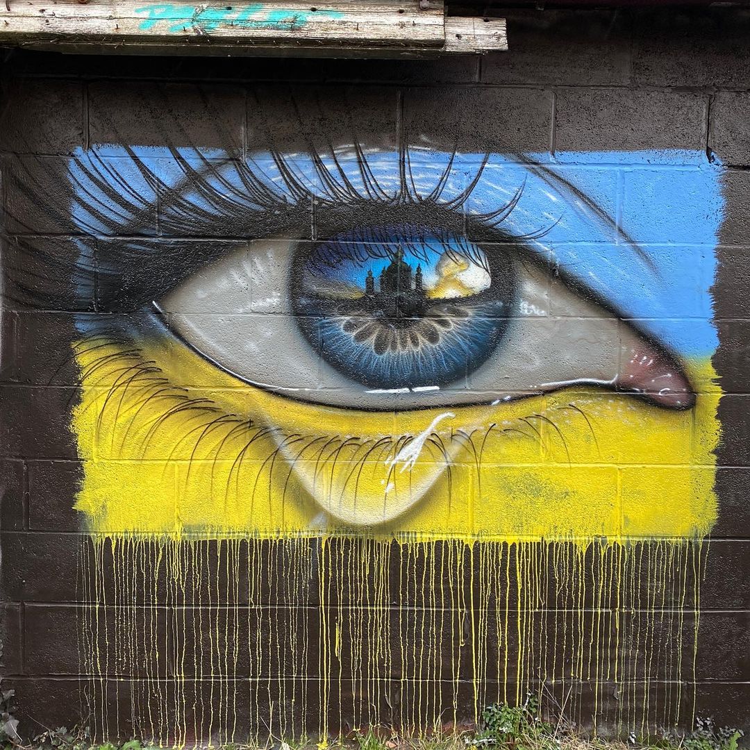 Screen shot of a mural of an eye shedding a tear/crying. The upper eyelid is painted in the blue of Ukraine's flag. The lower eyelid is painted in the yellow of Ukraine's flag. The mural was painted by the artist MyDogSighs.