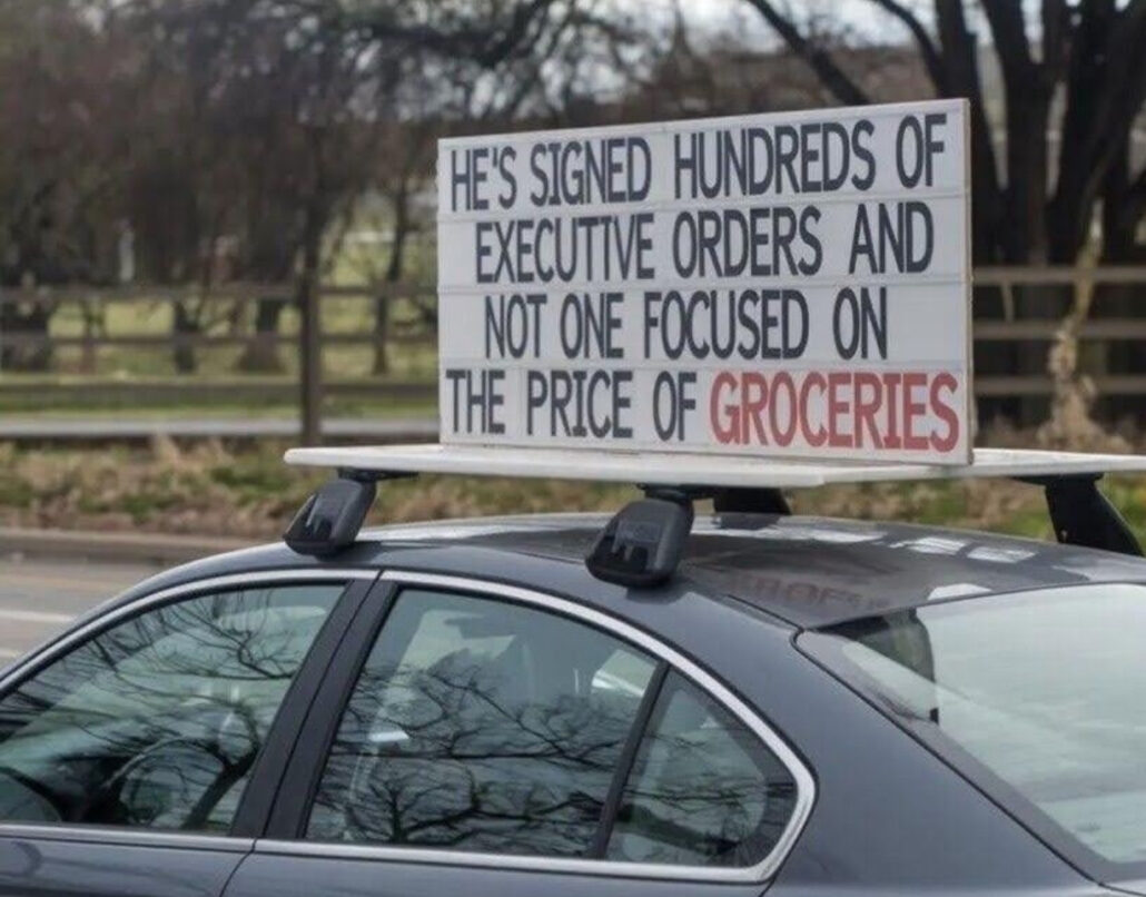 Car top sign reading "He's signed hundreds of executive orders and not one focused on the price of groceries."