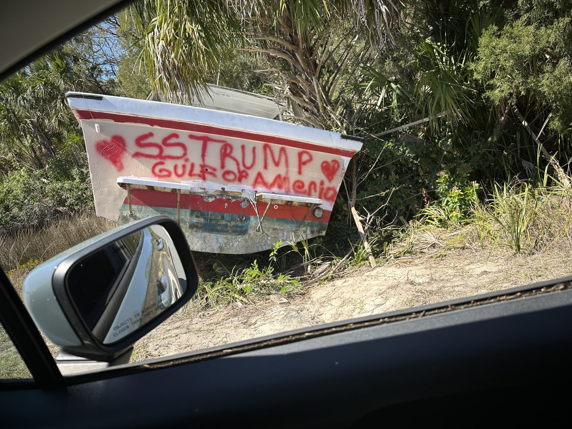 Wrecked boat on a roadside onto which someone has spray painted “SS TRUMP” and “Gulf of Ameria”