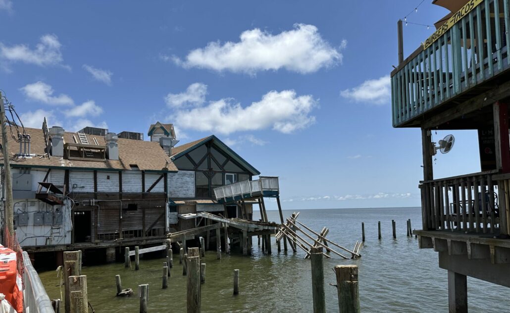 Empty piers in the water where a wooden building once stood. Adjacent buildings have visible damage. 
