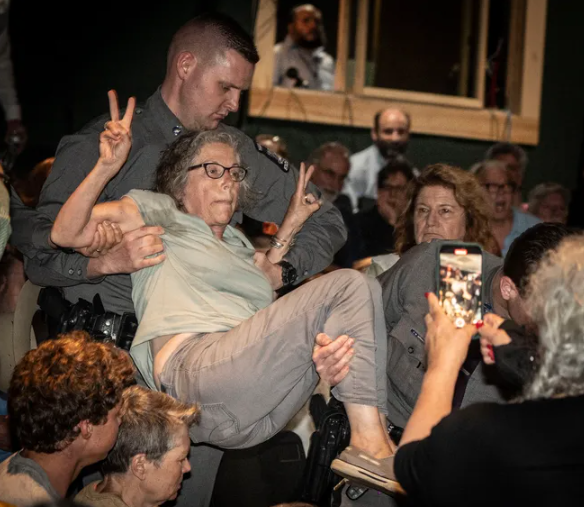 Photo of cops carrying a woman out of a town hall while she flashes peace signs.