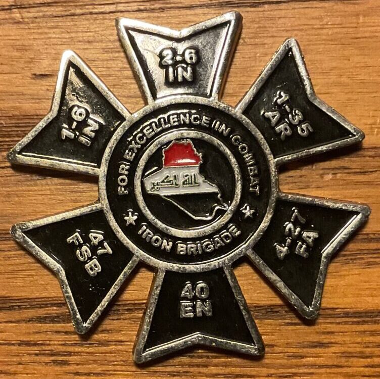 A US Army challenge coin. It is black and has 6 petals around a circular core. The circular core has an outline of Iraq with its flag superimposed on it. Above the flag is inscribed "For Excellence in Combat." Below the flag is inscribed "Iron Brigade. Each of the leaves has the name of one of the 2nd Brigade Combat Team/1st Armored Division. Each petal has the crest of each of the brigade's battalions: 1-6 Infantry, 2-6 Infantry, 1-35 AR, 4-27 Field Artillery, 40th Engineers, and the 47 Forward Support Battalion.