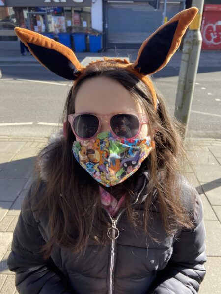 A white girl with long dark hair wearing pink sunglasses, a Pokemon-themed mask, and brown pointy Eevee ears, looks at the camera on a sunny urban street