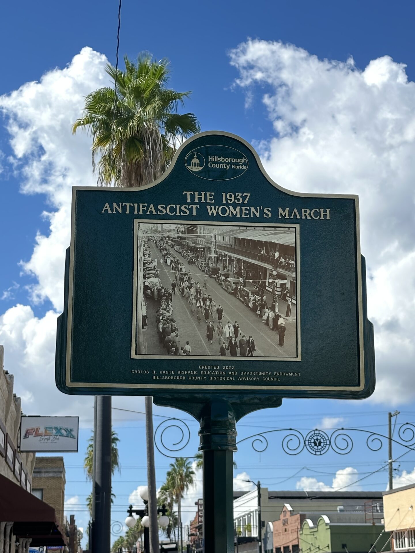 Historical marker with palm trees and a brilliant blue sky and puffy white clouds in the background.